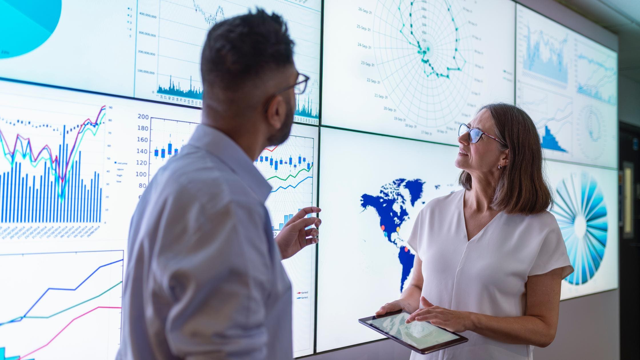 Two people having a discussion in front of large illuminated screens that are displaying graphs